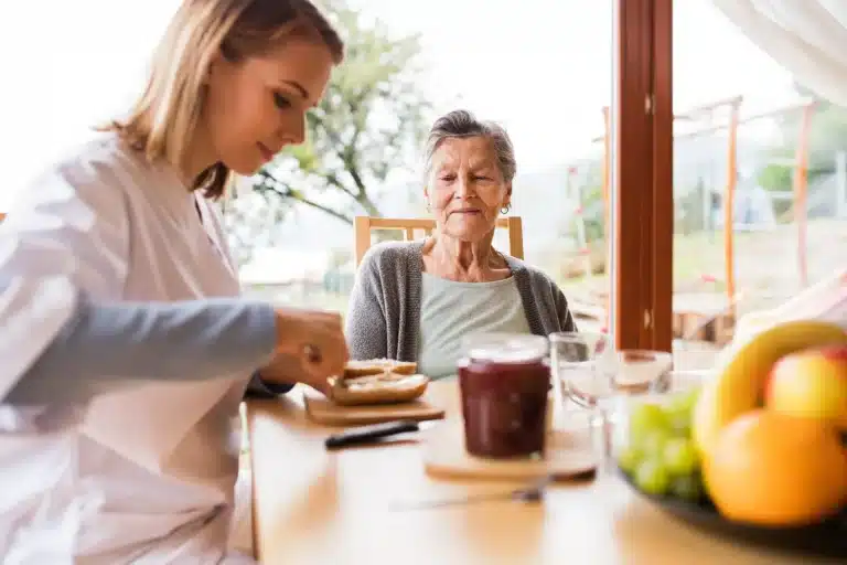 Caregiver helps an elderly woman at a sunlit dining table during breakfast, with toast, juice, and fruit visible nearby.